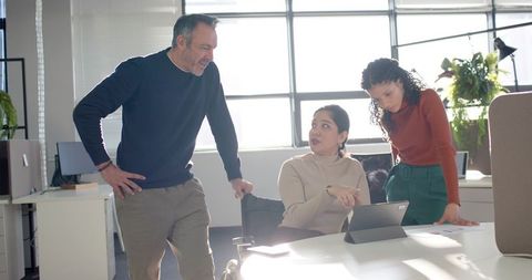 Diverse team collaborating around tablet in sunlit open-plan office, colleague pointing