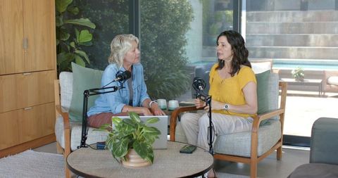 Two women recording podcast in modern living room casual interview with microphones and laptop