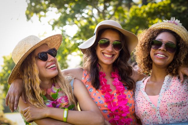 Three friends are standing together in a park on a sunny day, smiling and wearing sunglasses and hats. They are dressed in casual summer attire, enjoying each other's company. This image can be used for promoting outdoor activities, summer events, friendship themes, and leisure time.