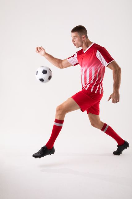 This image depicts a skilled Caucasian male soccer player in action, wearing a red uniform and kicking a soccer ball. Ideal for use in sports-related content, advertisements, training materials, and promotional campaigns for soccer events or athletic gear.