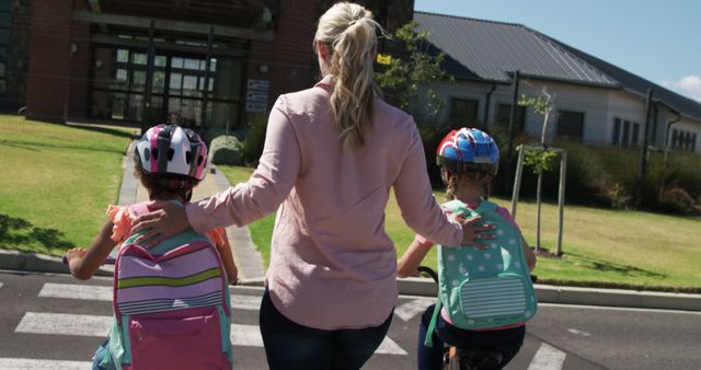 Mother accompanying two children carrying backpacks on their first day of school while using crosswalk with safety helmets on. Illustrates family bonding, child safety, and educational milestone. Useful for articles about back-to-school season, parenting advice, childhood education, and road safety awareness.