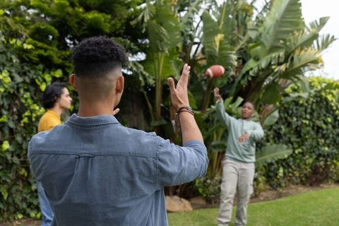 Young men tossing and catching football on backyard lawn with tropical hedge