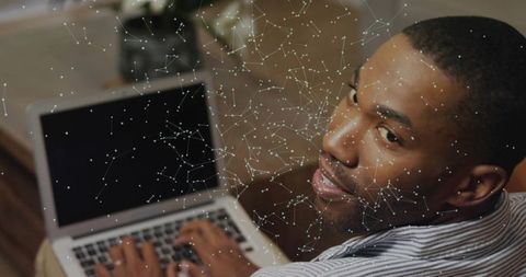 Man typing on laptop turning to camera with digital network overlay, cozy home workspace