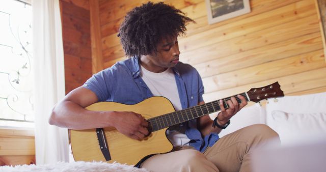Happy african american man sitting on sofa and playing guitar in log cabin, slow motion. Lifestyle, domestic life, countryside and nature concept.