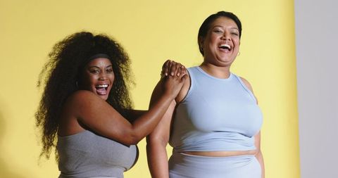 Joyful diverse women laughing in athletic wear on bright yellow background, body positive