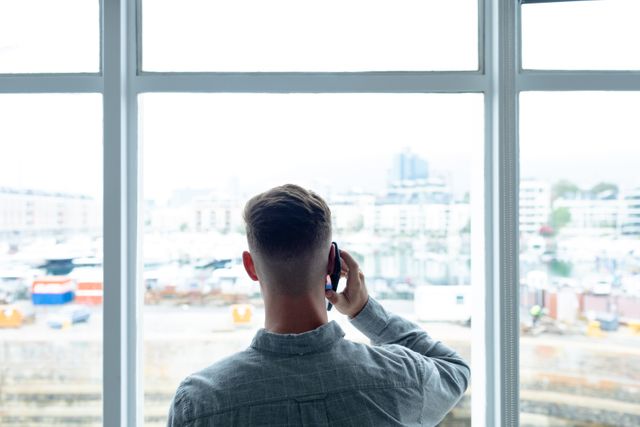 Rear view of a Caucasian businessman talking on a mobile phone near a large window in a modern office. The cityscape outside the window suggests an urban setting. This image can be used for business, communication, technology, and corporate themes, illustrating modern work environments and professional interactions.