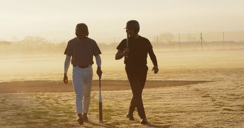 Diverse baseball teammates walking across dew-covered field at sunrise with bats and helmets