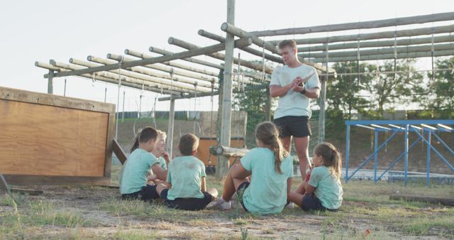 Group of children sitting on the ground attentively listening to their coach. They are gathered in an outdoor obstacle course area. This image is ideal for promoting summer camps, team-building activities, or physical education programs. It emphasizes learning, guidance, and the importance of teamwork in children's development.