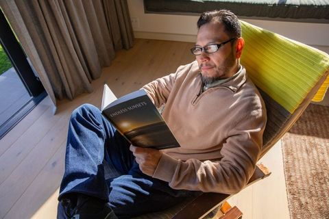 Asian man reading endless sunsets in sunlit lounge chair, relaxing at home with glasses