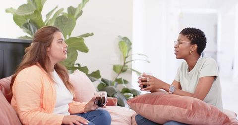 Two friends chatting on plush sofa with coffee mugs and velvet pillows in cozy living room