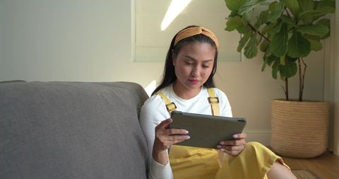 Sunlit asian woman using tablet on grey sofa wearing yellow overalls and headband