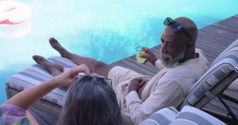 Senior couple relaxing poolside on striped loungers, enjoying cocktail and sunglasses