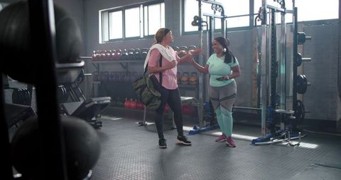 Diverse women chatting in sunlit gym wearing activewear carrying duffel bag and smartphone