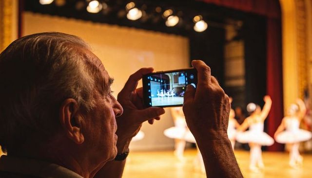 Senior man filming ballet recital on smartphone in golden theatre lighting