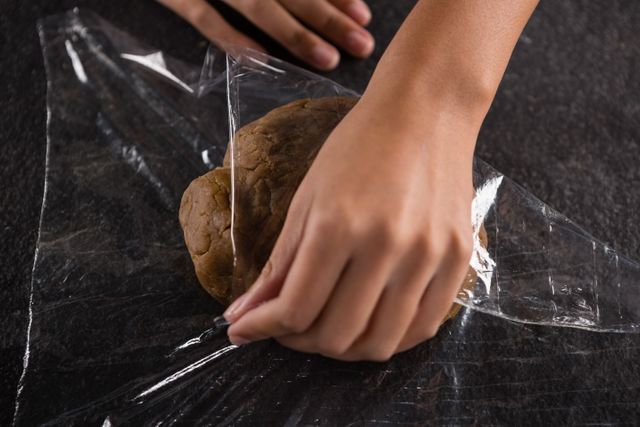 Hands wrapping dough in plastic wrap on a dark surface. Perfect for illustrating baking preparation, kitchen activities, and homemade cooking processes. Ideal for use in food blogs, recipe websites, and culinary magazines.