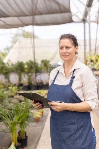 Greenhouse gardener inspecting potted plants with clipboard and pen wearing denim apron