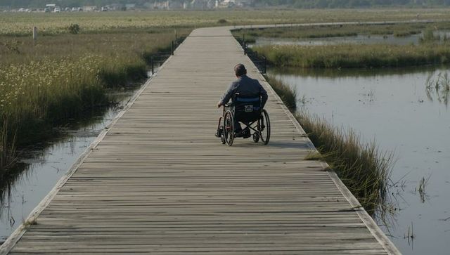 Man rolling wheelchair along wooden boardwalk through marsh toward coastline