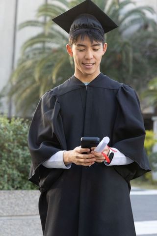 Asian male graduate wearing mortarboard and gown holding diploma and smartphone outdoors