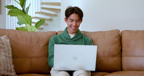 Smiling Man in Green Hoodie Using Laptop on Leather Couch