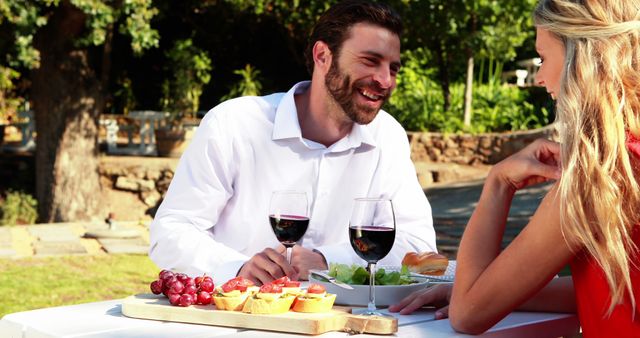 A young Caucasian couple enjoys a romantic outdoor meal with wine and a cheese platter, with copy space. Their cheerful engagement and the intimate setting suggest a special occasion or celebration.