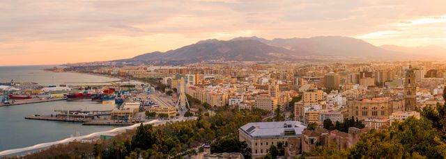 Panoramic view of Malaga cityscape at sunset showing harbor, distant mountains, and urban buildings under soft evening light. Ideal for travel agencies, tourism brochures, city travel guides, and websites promoting Mediterranean destinations.