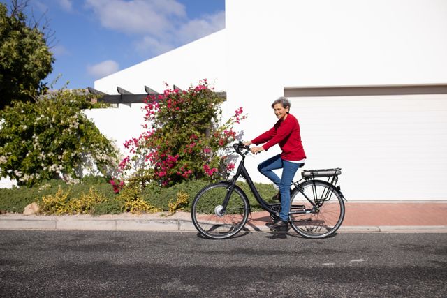 Side view  of a senior Caucasian woman with short grey hair wearing a red sweater riding a bicycle in the street, and smiling in the sun.