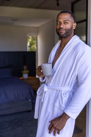 Mature african american man in white bathrobe holding coffee mug in minimalist bedroom