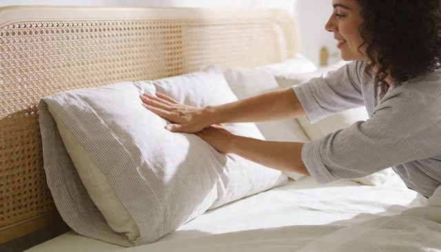 Woman smoothing pillow on sunlit bed with cane headboard and soft cozy neutral linens