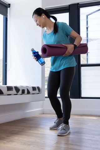 Mature woman holding yoga mat and water bottle preparing for home workout in bright studio
