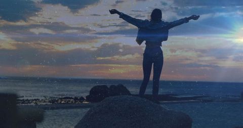 Silhouette woman standing on coastal boulder stretching arms at sunset with sun flare