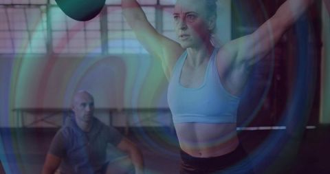 Woman pressing kettlebell overhead while coach observing in moody gym light