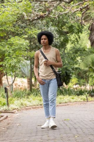 African american man walking in leafy park holding coffee and wearing crossbody bag