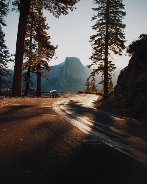 Depicting a serene mountain road with a car surrounded by sunlit trees and a distant mountain. Ideal for use in travel advertising, road trip promotions, nature documentaries, or any content related to scenic drives and exploration.