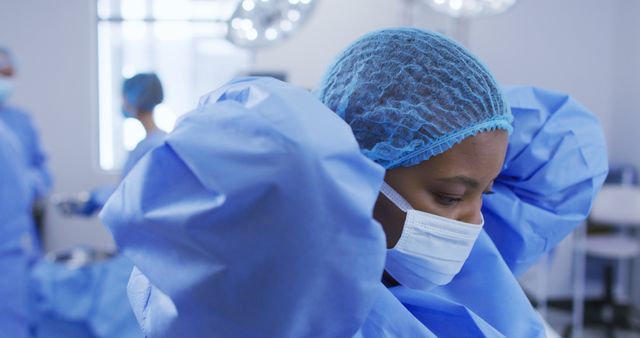 African american female surgeon wearing surgical cap, putting face mask on in operating theatre. medicine, health and healthcare services during covid 19 coronavirus pandemic.