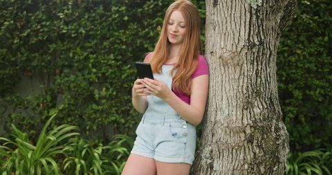 Red-haired woman leaning on tree trunk using smartphone in backyard garden, casual lifestyle
