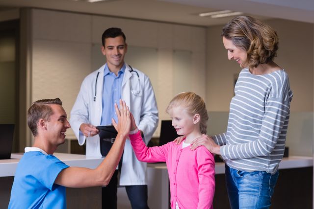 Doctor giving high five to young girl in hospital corridor while nurse and mother look on. Ideal for use in healthcare, pediatric care, family health, and medical professional promotional materials.