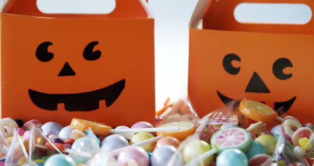 Two orange Halloween-themed boxes with jack-o'-lantern faces are surrounded by an assortment of colorful candies, with copy space. The festive containers and sweets evoke the spirit of Halloween celebrations and trick-or-treating traditions.