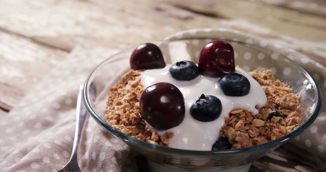 Perfect for promoting healthy eating and breakfast ideas. Picture shows a clear glass bowl filled with crunchy granola topped with creamy yogurt. Fresh cherries and blueberries add color and nutritional value. Background features a rustic wooden table and a fabric napkin, adding a homely and inviting feel. Ideal for food blogs, nutrition articles, and breakfast-themed advertisements.