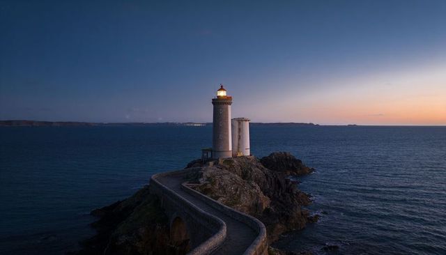 Lighthouse beacon at dusk on rocky promontory with curving causeway and lantern light