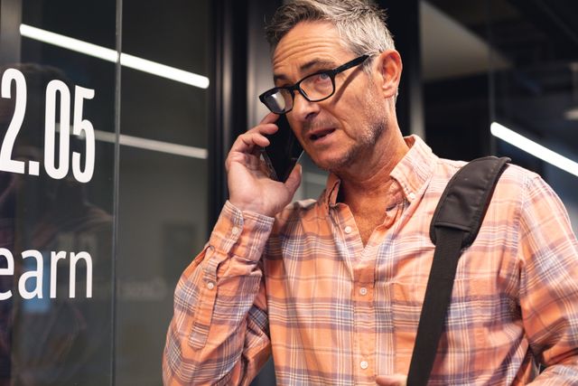 Middle-aged Caucasian businessman talking on smartphone while holding a bag in an office. He is wearing a casual plaid shirt and glasses, appearing engaged in conversation. Useful for illustrating business communication, corporate environments, professional interactions, and modern office settings.