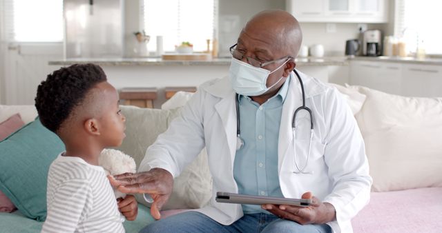 A healthcare professional is examining a young boy at home. The pediatrician is using a tablet and wearing a protective mask, showcasing a contemporary, safety-first approach to medical care. This setup emphasizes the consideration for health during home visits. Suitable for use in articles promoting home healthcare, medical professions, child healthcare, and the role of technology in modern medical practices.