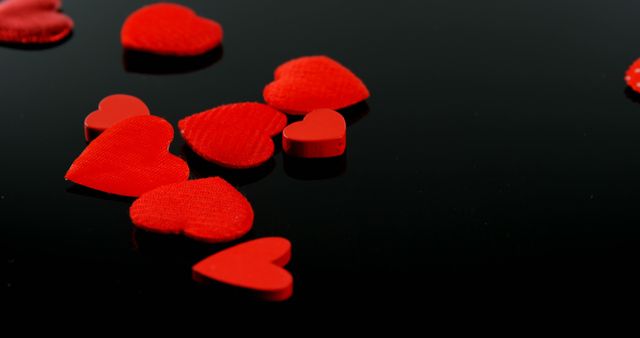 Red fabric hearts are scattered across a reflective black surface, with copy space. Their vibrant color and heart shape symbolize love and romance, often associated with Valentine's Day.