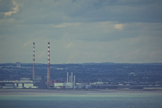 Panoramic view of industrial chimneys releasing smoke under a cloudy sky. Coastal area with diverse industry architectures. Perfect for illustrating environmental issues, industrial activities, pollution awareness, and urban landscapes.