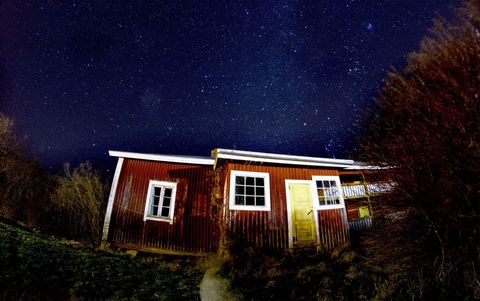 Rustic red cabin standing beneath expansive starry night sky with glowing yellow door