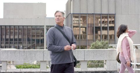 Mature man walking and woman strolling on city walkway with smartphones by stone railing