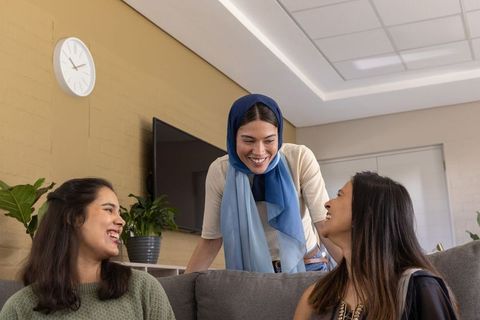 Young women chatting on sofa, woman wearing blue headscarf leaning in and laughing now