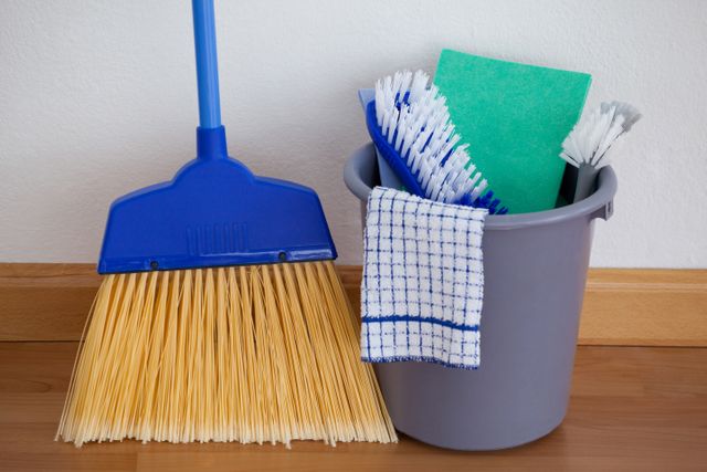 Various cleaning tools including a broom, bucket, brushes, sponge, and cloth on a wooden floor against a wall. Ideal for illustrating household chores, cleaning supplies, and hygiene practices. Suitable for articles, advertisements, and blogs related to home cleaning and maintenance.
