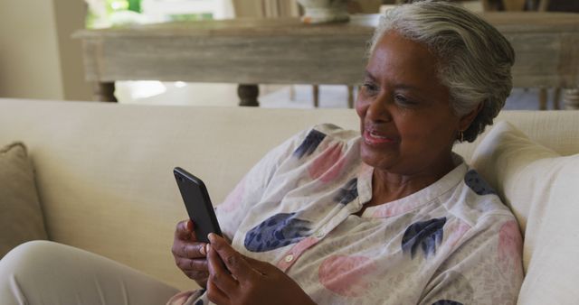 Elderly woman enjoys using a smartphone while sitting on a couch at home. Great for use in advertising technology for seniors, promoting senior-friendly apps, or addressing elderly lifestyle themes such as staying connected or digital literacy.