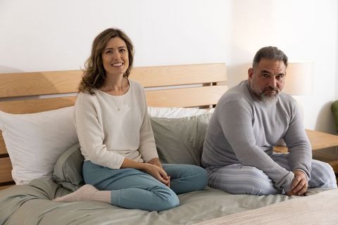 Couple sitting on bed sharing calm morning, smiling woman and contemplative man in pajamas