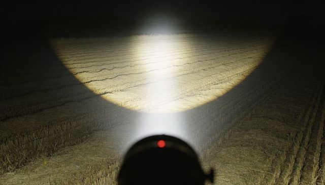 Handheld flashlight illuminating harvested field furrows at night with red indicator glow